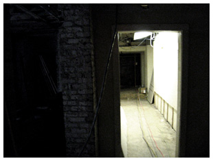 A dark image of a basement and a hallway.
Construction debris is littered on the floor.
There are extension cords and temporary lighting in place.